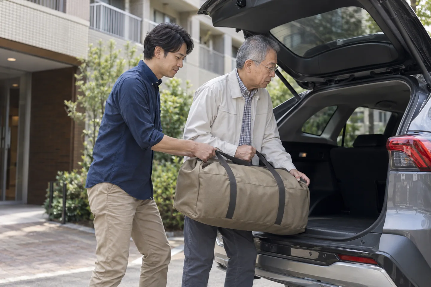 Two adult men lifting a plain travel bag into a car outside an apartment
