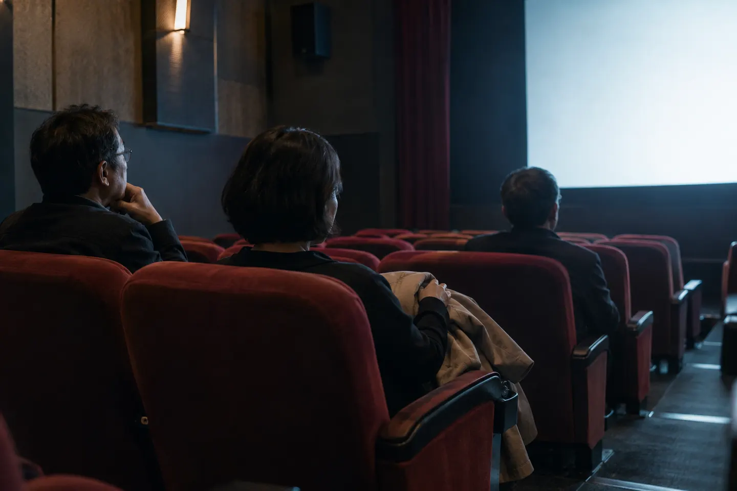 Três adultos sentados entre poltronas vermelhas de cinema junto à luz de uma tela em branco
