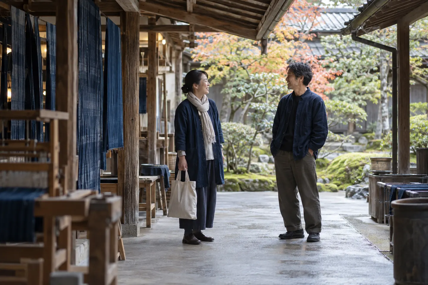 Two adults talking under a covered weaving-workshop courtyard beside hanging indigo fabric and a moss garden